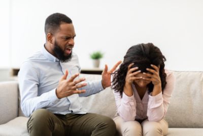 A couple sitting on a couch where the man is speaking angrily to a woman with her head in her hands