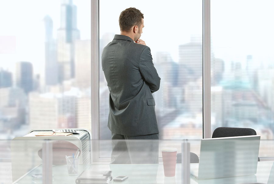 Professional standing alone in an office looking out the window while reflecting after a stressful work experience, representing trauma triggers and workplace anxiety.